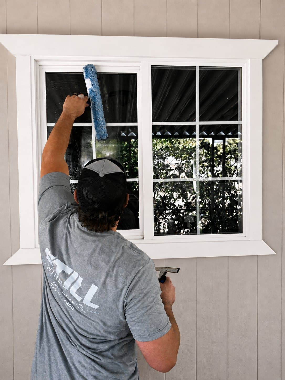 Still Window Co washing the upper panes of a divided-light residential window, applicator in one hand and squeegee in the other.